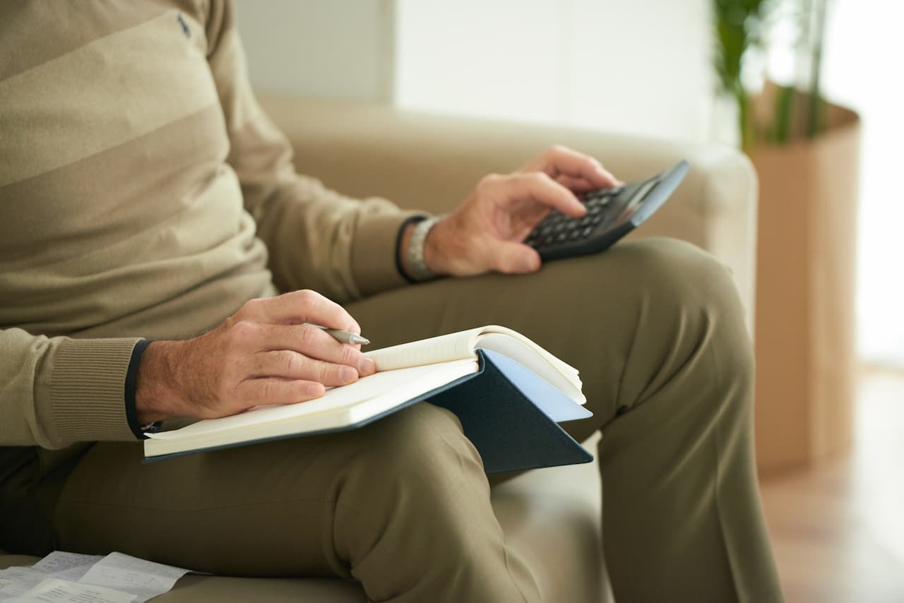 Homem sentado em um sofá com um caderno de notas apoiado na coxa direita, e sua mão está sobre o caderno, enquanto segura uma caneta. Na mão esquerda ele segura uma calculadora, apoiando-se na coxa esquerda.