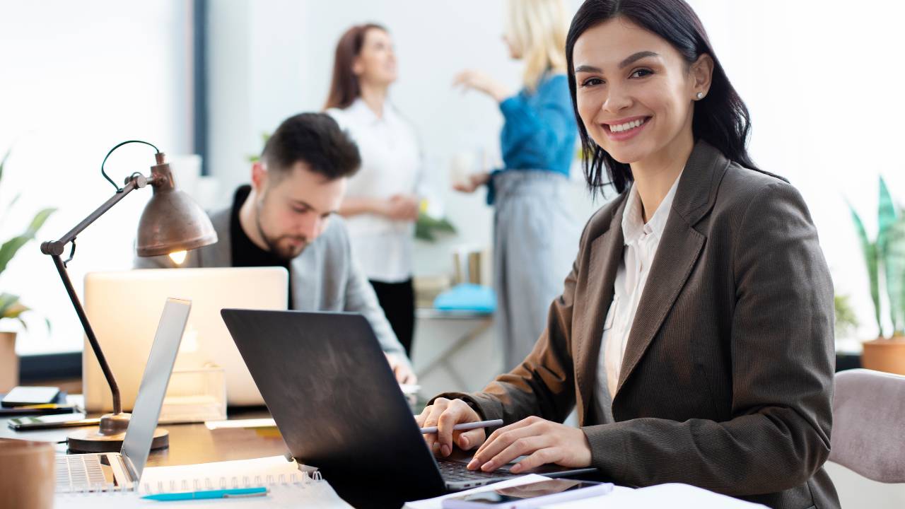Pessoas felizes com seus empregos trabalhando em um escritório. O foco da imagem está em uma mulher jovem sorridente, digitando em um notebook.