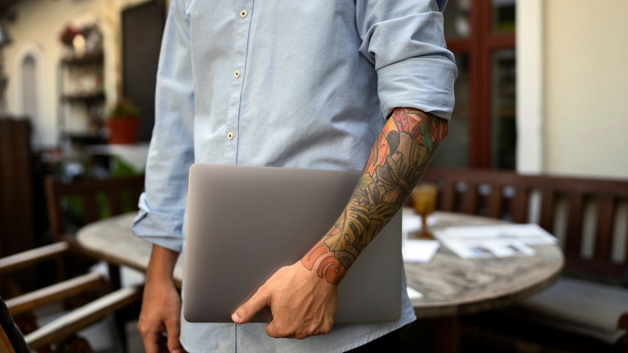 Pessoa vestida formalmente, em pé, segurando um notebook fechado com o braço esquerdo, o qual possui muitas tatuagens.