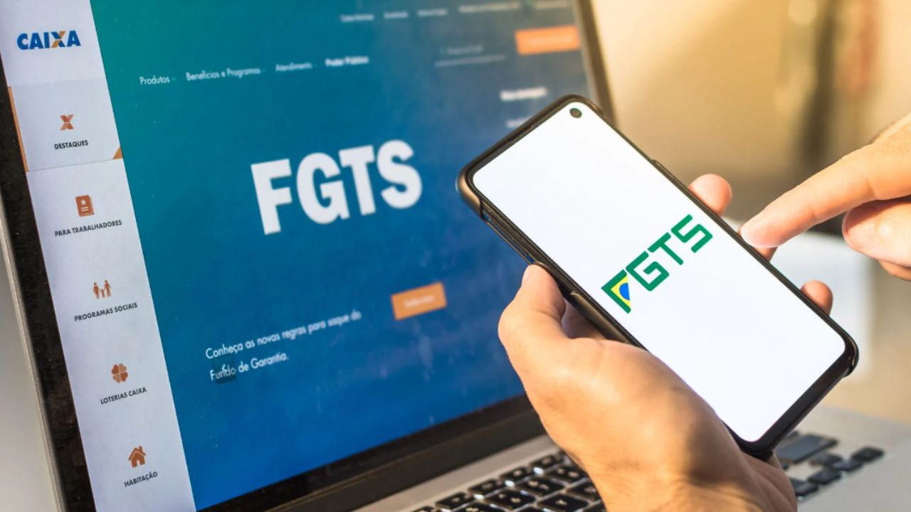 Close up em mão segurando celular com aplicativo do FGTS aberto. Ao fundo há um notebook no site do FGTS.