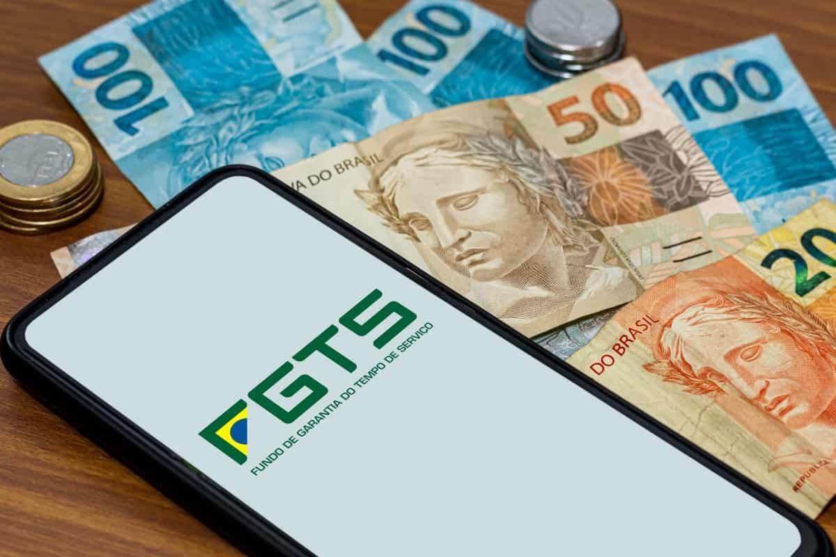 FGTS. Celular com o app do FGTS aberto, em cima de uma mesa com diversas notas e moedas de dinheiro.