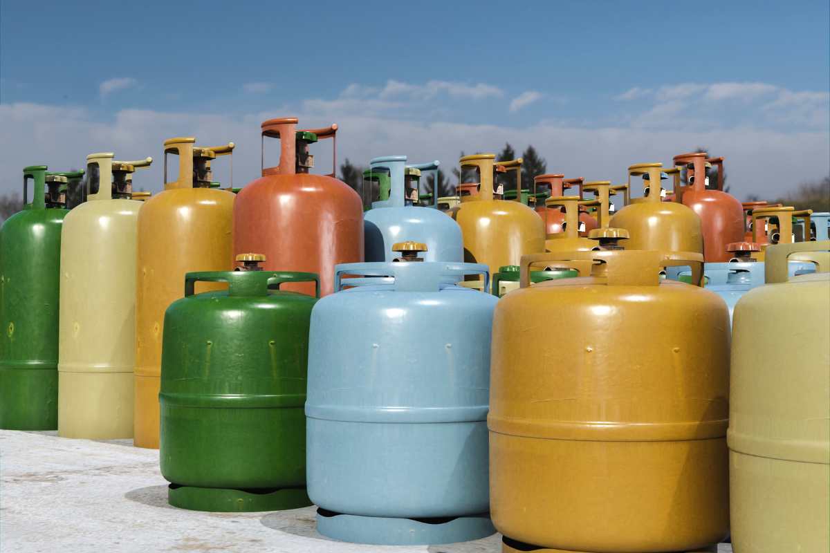 Gás. Diversos cilindros de gás de cozinhas, de diferentes cores, dispostos em fila a céu aberto.
