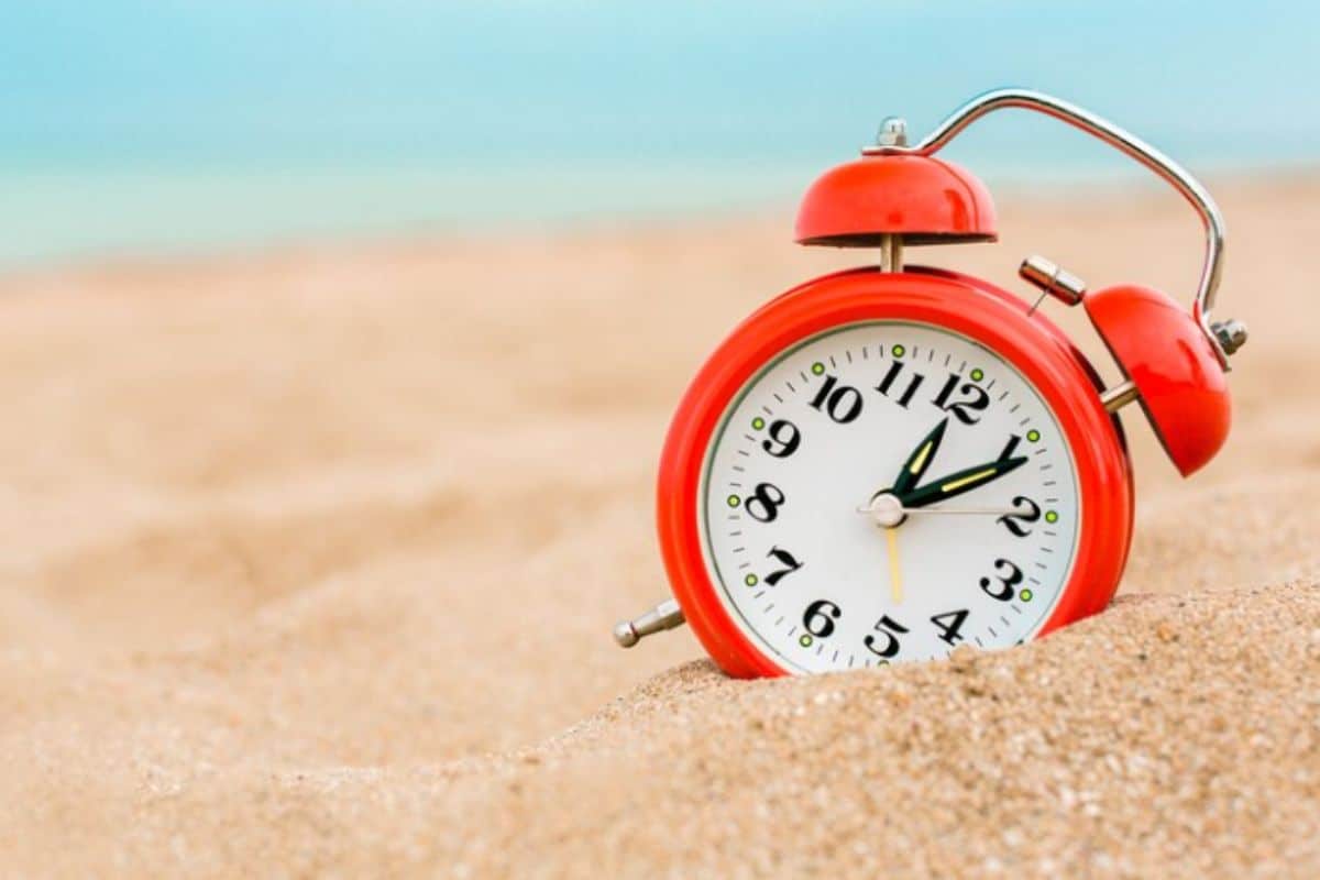Horário de Verão. Relógio despertador parcialmente enterrado em areai da praia em um dia ensolarado, fazendo alusão ao horário de verão.