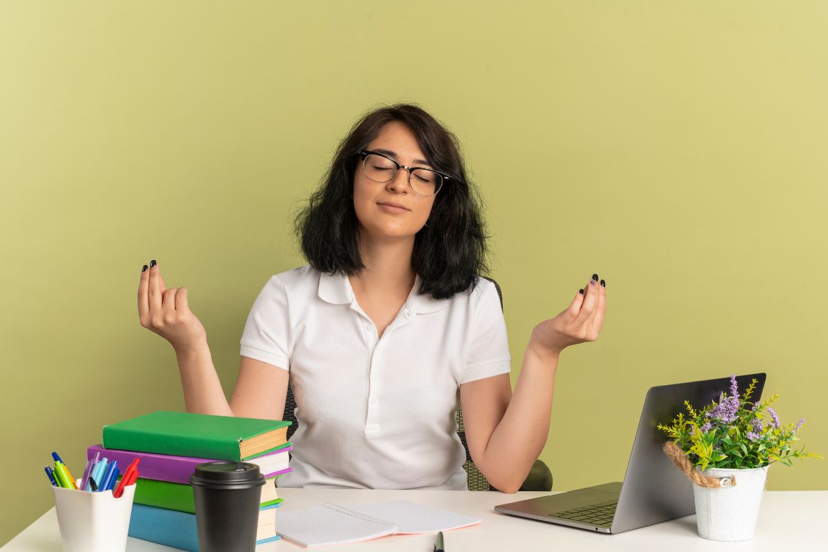 Mulher sentada em escritório, com uma mesa cheia de cadernos, canetas e etc. Para promover saúde mental no trabalho, ela está usando a técnica de meditação.
