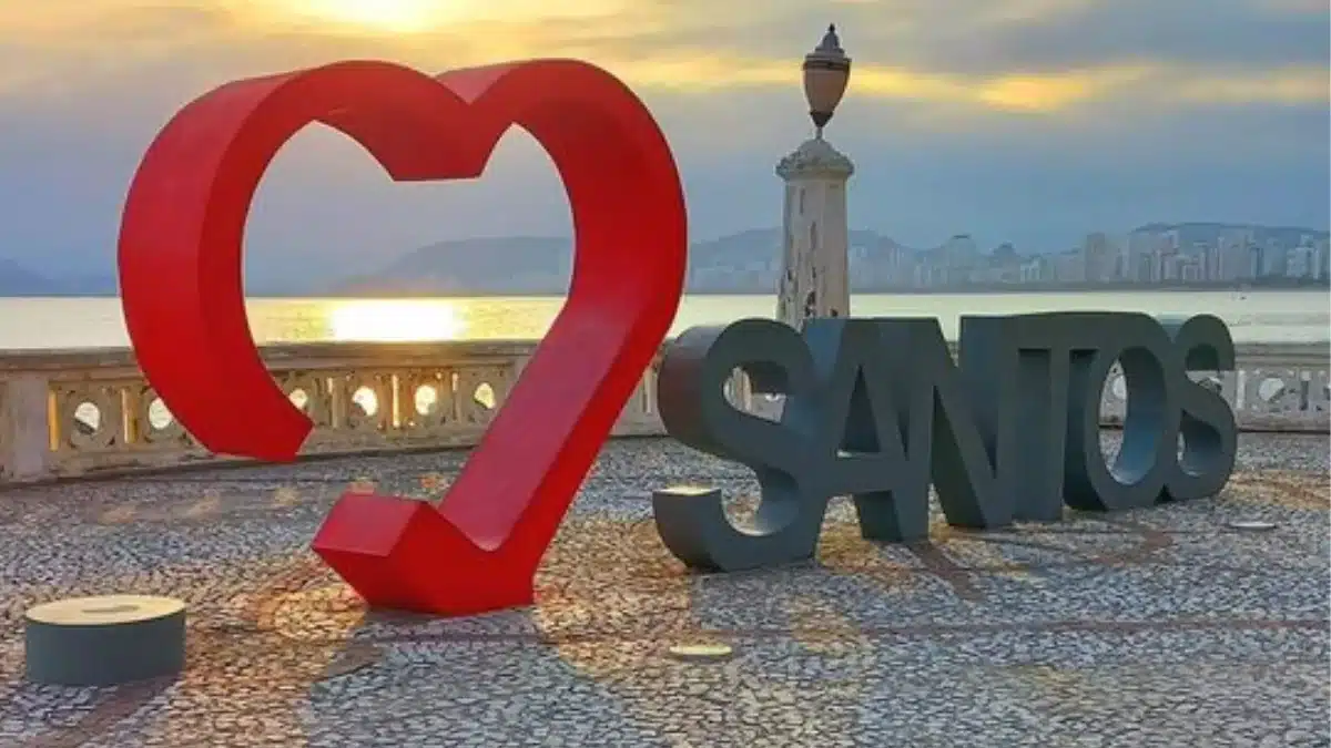 Escultura de coração ao lado de "Santos" em homenagem à cidade litorânea. Ao fundo vê-se o mar e o pôr do sol.