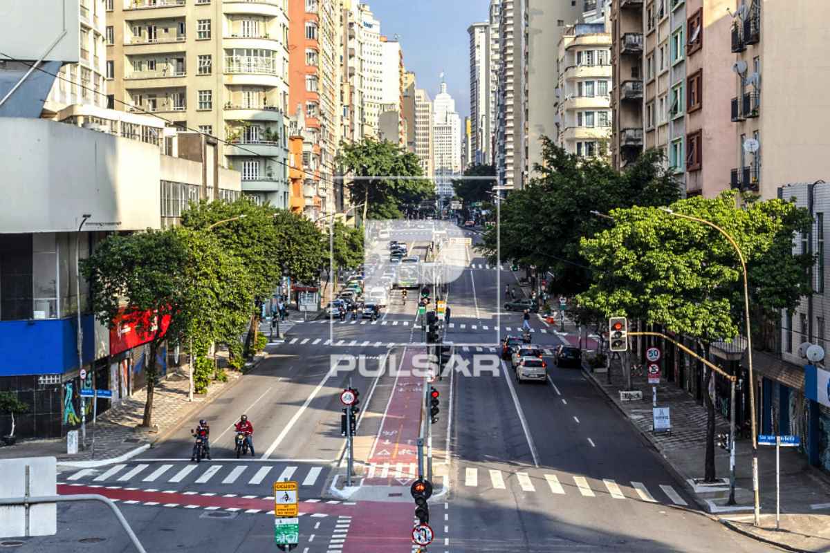 Vista de cima da Avenida São João, no centro de São Paulo. Vê-se árvores, muitos prédios, motociclistas e carros.