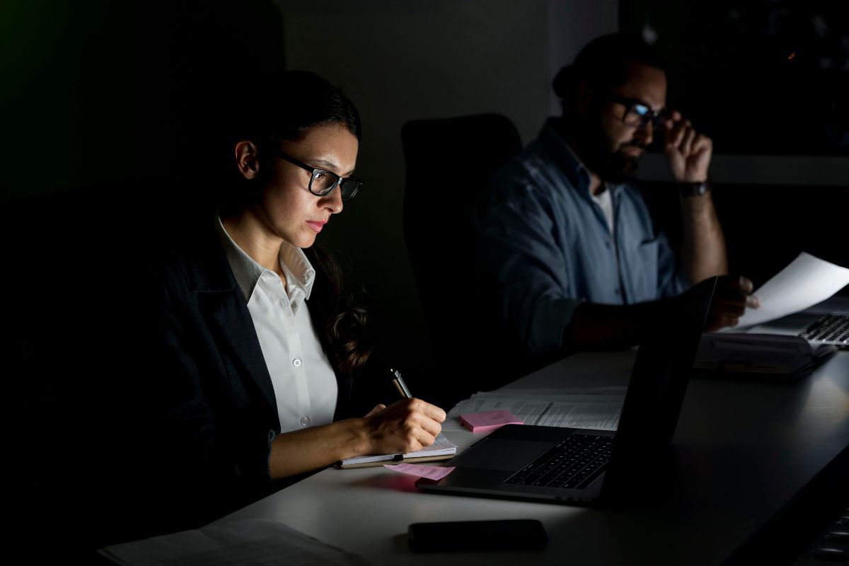 Dois funcionários em um escritório escuro, usando computadores e fazendo anotações durante trabalho noturno.