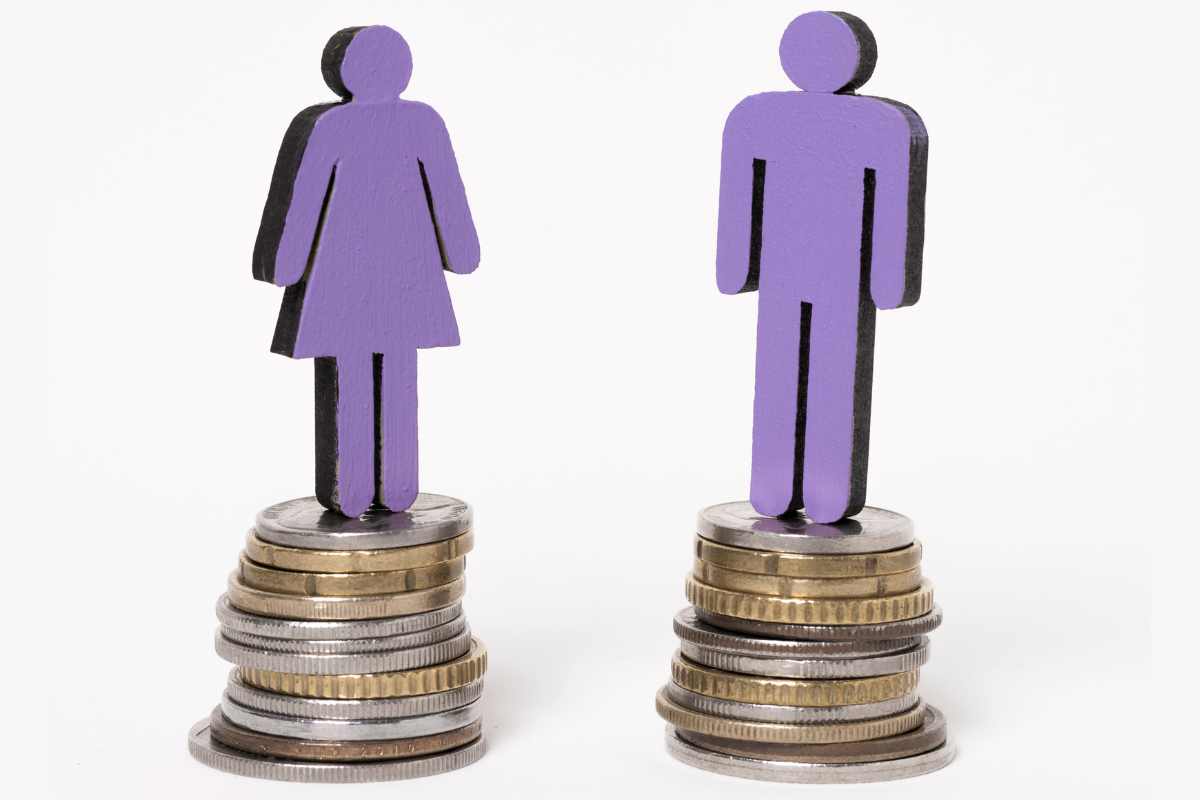 Boneco feminino e masculino em cima de pilhas de moedas em lados opostos, em alusão à diferença salarial entre homens e mulheres.