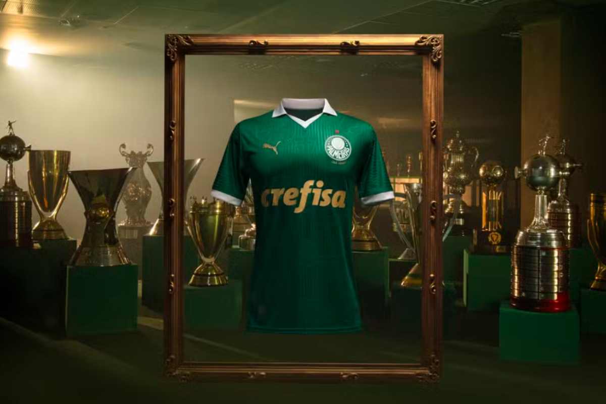 Banner de divulgação da nova camisa do Palmeiras. Ela está centralizada na imagem, com uma moldura de quadro de madeira ao seu redor. Ao fundo, há diversos troféus.