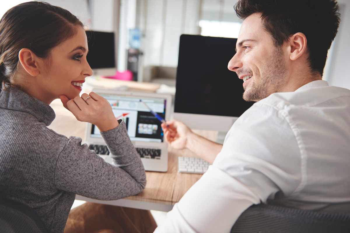 Em um escritório, uma mulher e um homem sorridentes estão sentado à mesa, bem próximos, em um íntimo relacionamento no trabalho.
